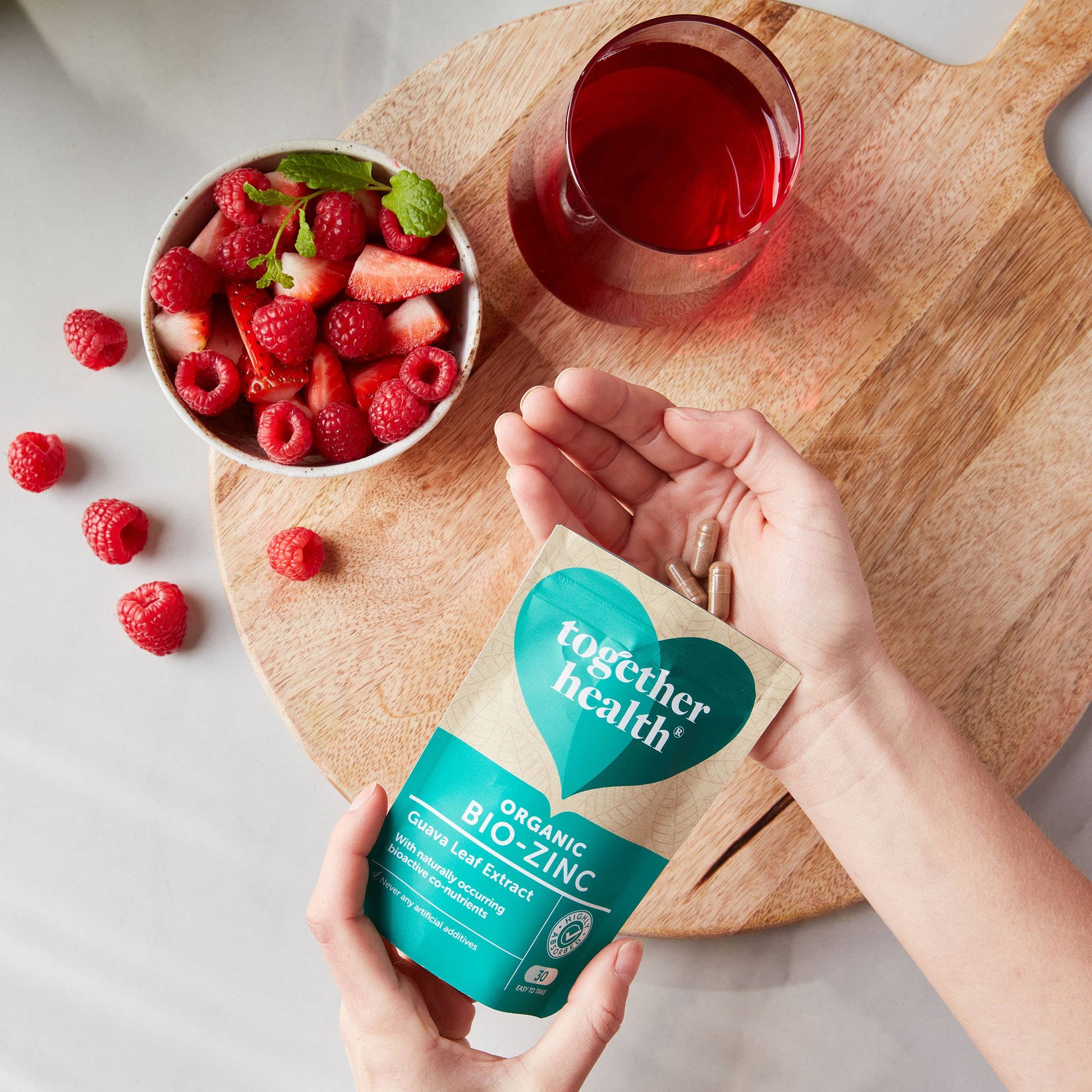 A hand holds a pouch of organic guava leaf supplements, surrounded by fresh berries, a mint garnish, and a glass of red beverage.