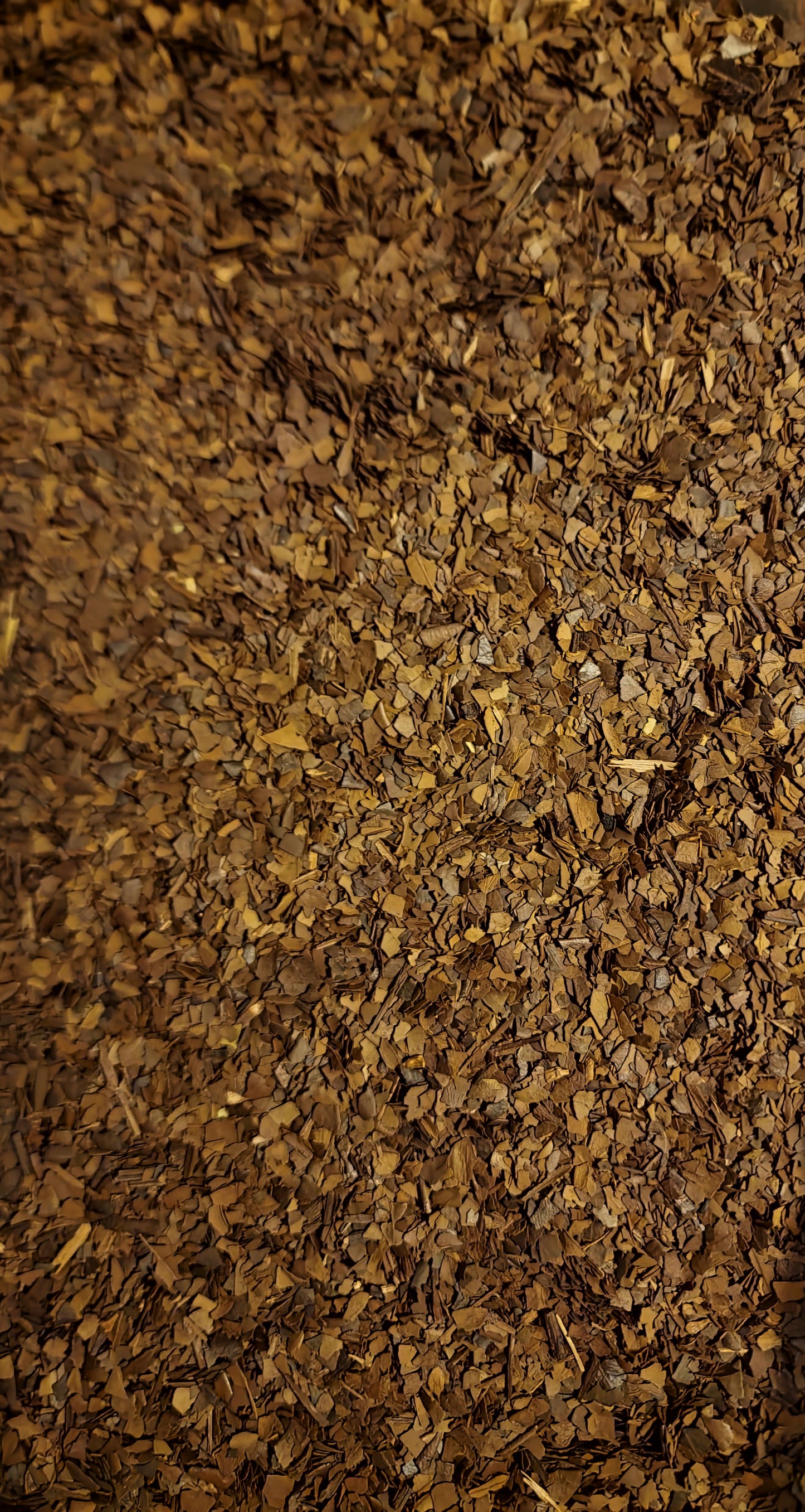 An overhead macro photograph of loose Apothecary Grade Roasted Yerba Mate, displaying the rich uniform mixture of toasted charcoal-brown, bronze, and tan cut leaves and stems of Brazilian Ilex paraguariensis.