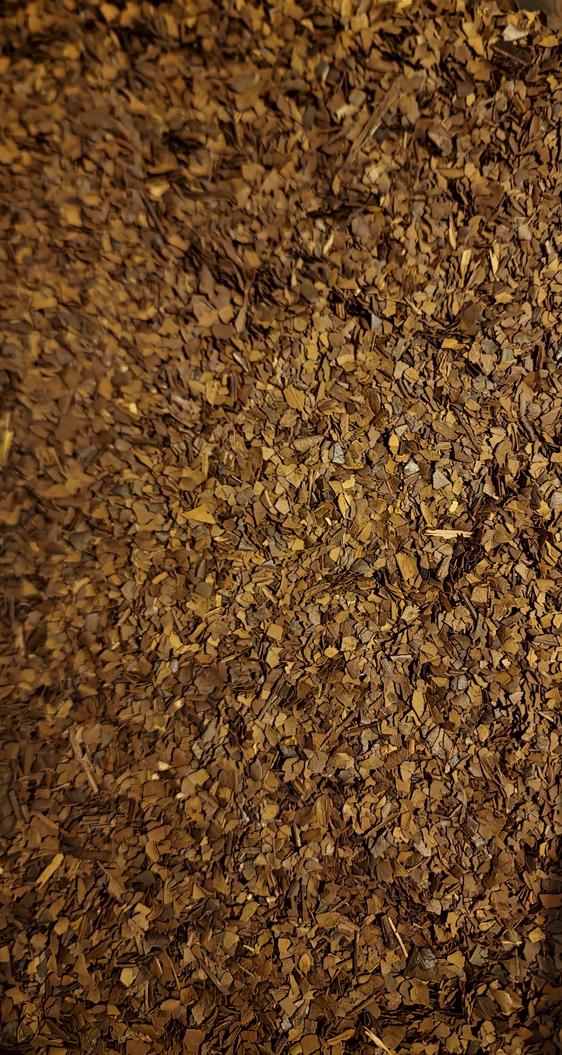 An overhead macro photograph of loose Apothecary Grade Roasted Yerba Mate, displaying the rich uniform mixture of toasted charcoal-brown, bronze, and tan cut leaves and stems of Brazilian Ilex paraguariensis.